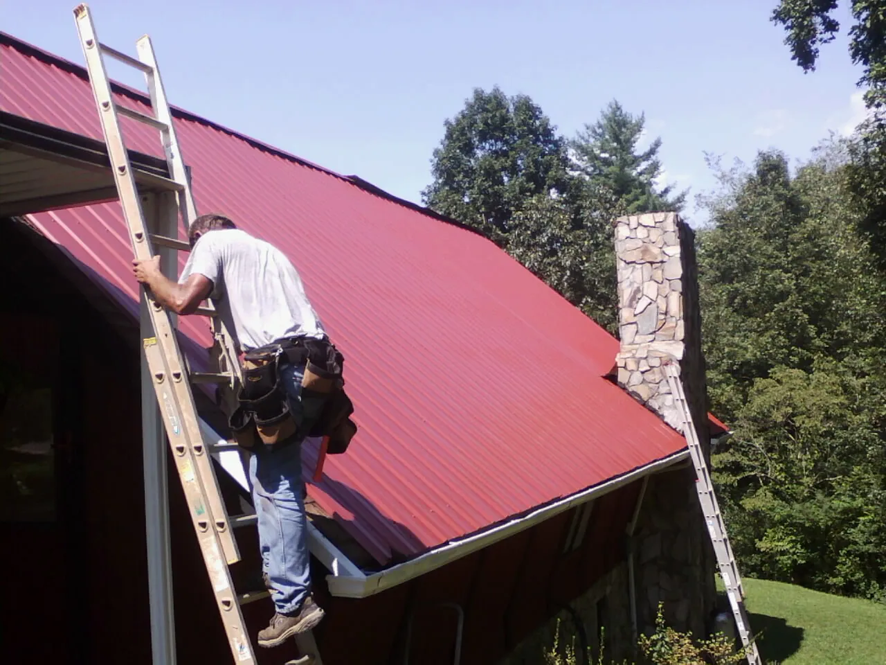 Roofer on a ladder beside a red metal roof and stone chimney — Soffit & Fascia Repair by Roofing Brothers in Cawood